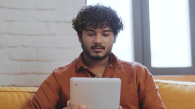 Kıvırcık saçlı genç bir adam yumuşak bir kanepede otururken tabletine odaklanır. Güneş ışığı pencereden süzülüp sıcak ve davetkar bir atmosfer yaratır..