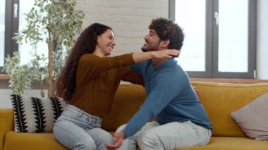 Bir çift, parlak bir oturma odasındaki sarı bir kanepede neşeli bir anı paylaşıyor. Gülüyorlar ve birbirlerine uzanıyorlar, sıcak, samimi bir atmosfer yaratıyorlar. Güneş ışığı boşluğu doldurur.