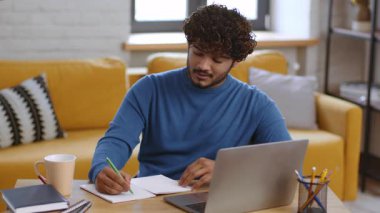 Bir adam, rahat bir oturma odasında, ahşap bir masada oturmuş deftere bir şeyler yazıyor. Yakınlarda bir dizüstü bilgisayarı açık ve bir fincan kahvesi var. Düşünceli bir ifadeyle işine odaklanmış..
