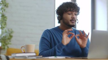 Kıvırcık saçlı bir erkek ahşap bir masada oturuyor ve aktif olarak sanal bir toplantıya katılıyor. Kulaklık takar ve konuşurken el kol hareketi yapar..