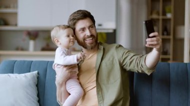 Bir baba ve bebeği kanepeye oturup selfie çekerken geniş bir gülümseme sergilerler. Sıcak oturma odasında yumuşak bir ışık ve sıcak bir atmosfer var. Neşeli bir aile anını yakalıyor..