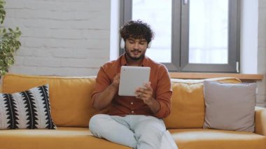 Genç bir erkek sarı bir kanepede rahatça oturuyor, tablet kullanıyor ve içeride vakit geçiriyor. Parlak odada geniş pencereler ve rahatlatıcı bir atmosfer var..