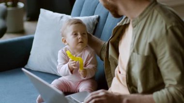 Aile ortamında kanepede oturan ve elinde muz tutan bir bebek görülüyor. Ebeveyn bebeğin yanında, sıcak bir atmosferi olan bir dizüstü bilgisayarda çalışıyor, neşeli bir deneyim yaratıyor..