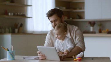 Neşeli bir baba bebeğiyle mutfak masasında oturuyor, ikisi de tabletle meşgul. Güneş ışığı pencereden akar ve sıcak evlerinde sıcak bir atmosfer yaratır..