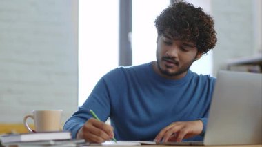 Kıvırcık saçlı genç bir adam ahşap bir masada oturur, dizüstü bilgisayarına odaklanırken notlar yazar. Yakınlarda bir kahve fincanı var ve arkasından doğal ışık geliyor..
