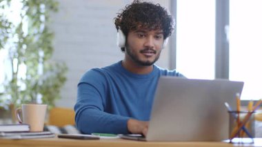 Kıvırcık saçlı genç bir adam kulaklık takarken çağdaş bir çalışma alanında dizüstü bilgisayar kullanıyor. Yanında bir kupa ve kırtasiye malzemesi ile işiyle meşgul görünüyor. Doğal ışık odayı dolduruyor..