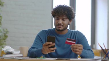 Genç bir adam iyi aydınlatılmış bir odada ahşap bir masada oturuyor. Bir elinde akıllı telefon, diğer elinde kredi kartı var. Çevrimiçi bir aktiviteye katılmış. Not defterleri ve kahve fincanı yakınlarda..