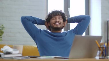 Gülümseyen bir erkek, ellerini başının arkasına koyup, rahat bir şekilde modern bir çalışma alanında oturup rahatlamanın tadını çıkarır. Doğal ışık geniş pencerelerden içeri akar ve sıcak bir atmosfer yaratır..