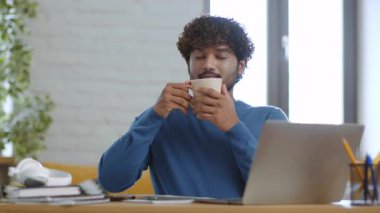 Genç bir erkek bir masada oturuyor, bir fincan kahvenin tadını çıkarıyor. Doğal ışık odayı doldurur ve sıcak bir atmosfer yaratır. Kitaplar ve dizüstü bilgisayarlar yakınlarda, bir çalışma oturumunu gösteriyor..