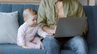 Meraklı bebekleri yakınlarda oynarken bir ebeveyn mavi bir kanepede dizüstü bilgisayarla oturuyor. Konforlu ev ortamı, teknolojiyle rahat bir bağlantı ve öğrenme anı gösteriyor..