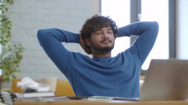 Kıvırcık saçlı genç bir adam masada oturuyor, elleri başının arkasında arkana yaslanıyor. Yakınlarda bir dizüstü bilgisayar ve bitkilerle aydınlık bir odada rahat görünüyor..