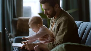 Bir baba sıcak bir kanepede oturmuş bebeğini tutuyor. Bebek dizüstü bilgisayara uzanırken baba dikkatlice etkileşime giriyor. Oda yumuşak doğal ışıkla aydınlık..