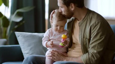 Babam parlak bir oturma odasında renkli bir oyuncakla oynarken bebeğin başını nazikçe öper. Sahne, doğal ışıkla vurgulanan sıcaklık ve sevgiyle dolu bir bağ anı gösteriyor..