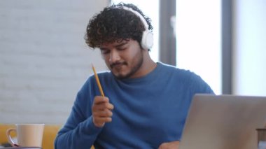 Genç bir erkek bir masada oturuyor, derslerine odaklanmış. Kulaklık takıyor ve not almak için kalem kullanıyor. Yumuşak ışık odada sıcak bir atmosfer yaratır..