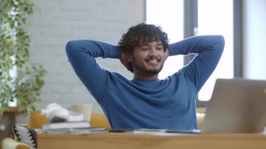 Kıvırcık saçlı bir erkek rahat bir oturma odasındaki masada dizüstü bilgisayarına bakarken gülümsüyor. Güneş ışığı pencereden içeri akar ve sıcak bir ortam yaratır..