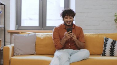 Neşeli genç bir erkek rahat sarı bir kanepede oturuyor, akıllı telefonuyla meşgul. Parlak odada sıcak ve davetkar bir atmosfer yaratan büyük pencereler var..