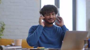 Mavi kazak giyen bir erkek profesyonel evde sanal bir toplantı yapıyor. Dizüstü bilgisayarına odaklanırken iki telefon kullanıyor. Doğal ışık odayı dolduruyor, sıcak bir atmosfer ekliyor..