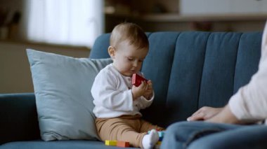 Bir bebek rahat mavi bir kanepede oturuyor, renkli bloklarla oynamaya odaklanmış. Hareketli ışık odaya akıyor, sıcak ve davetkar bir atmosfer yaratıyor..