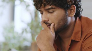 Kıvırcık saçlı bir erkek sıcak bir mekanda, düşüncelere dalmış bir şekilde oturur. Çenesini elinde tutar ve arka plandaki yeşil bitkilerin arasında derin bir şekilde yansıtır. Doğal ışık sahneyi aydınlatır.