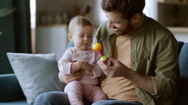 Bir baba bebeğini kucağında rahat bir kanepeye oturur. Baba, meraklı bebeğe renkli bir oyuncak gösterirken oyunla meşguldürler. Oda aydınlık ve davetkar..