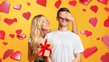 Sevgi dolu bir sevgili, elinde bir hediye tutarken erkek arkadaşının gözlerini gülümsemeyle kapatır. Kalpleri olan sarı bir zemin üzerinde duruyorlar, birlikte özel bir anı kutluyorlar..