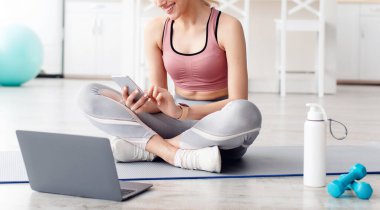 Sosyal ağlar, modern teknoloji ve evde eğitim için ekipman, yeni normal. Neşeli, genç, sarışın, spor kıyafetli bir kadın. Akıllı telefonuyla yazı yazıyor. Oturma odasında dizüstü bilgisayarla oturuyor.