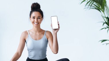 Bir kadın kapalı bir alanda bağdaş kurarak oturur. Gülümsüyor ve elinde bir akıllı telefon tutuyor. Arka planda yeşil bir bitki duruyor ve sahneye biraz renk katıyor..