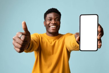 Sarılı yakışıklı Afro-Amerikan genç adam boş ekranlı modern cep telefonunu gösteriyor ve mavi stüdyo arka planında başparmağı var, en yeni mobil uygulamayı öneriyor.