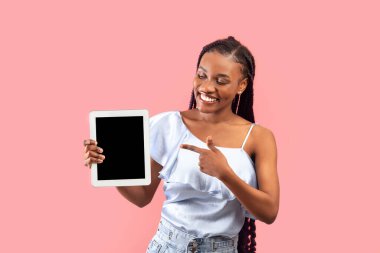 Çekici genç siyahi kadın pembe stüdyo arka planında boş ekranlı tablet, web sitesi için model veya yeni uygulama tasarımı gösteriyor. Milenyum Afrikalı Amerikalı kadın dokunmatik ped görüntüleme şablonu