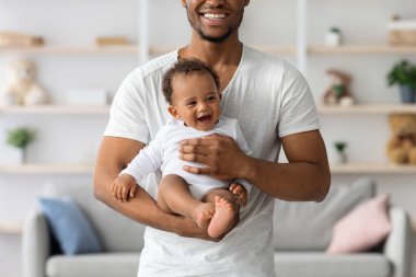 Gülümseyen siyah adam, elinde yeni doğmuş bebekle evde poz veriyor, neşeli Afrikalı Amerikalı bir babanın sevimli bir çocuğu kucağına alıp, babalığının tadını çıkartıyor, yakın çekim yapıyor.