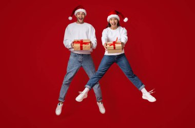 Sıcak süveter ve Noel Baba şapkaları giymiş neşeli bir çift havaya sıçrıyor, ellerinde parlak kırmızı bir arka planda süslü püslü hediyelerle..