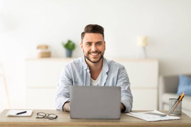 Başarılı bir serbest meslek sahibi. Mutlu adam iş yerinde dizüstü bilgisayarla oturuyor, gülümsüyor ve kameraya bakıyor, ev ofisindeki bilgisayarda uzaktan çalışıyor, boşluğu kopyalıyor. İnternet üzerinden uzak işten hoşlanan adam