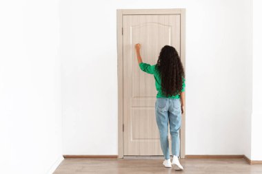 Bir dişi ziyaretçi kapalı bir ahşap kapıyı çalar ve içeri girme beklentisini gösterir. Parlak yeşil bir bluzu ve iyi ışıklandırılmış kapalı bir alanda kot pantolonu ile, günlük kıyafetleri içinde tam boy ayakta duruyor..