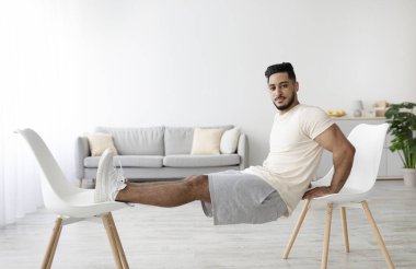 Atletik genç Arap adam evde iki sandalyeyle güç egzersizi yapıyor. Milenyum Ortadoğu 'lu adam kol kaslarını çalıştırıyor, iç organları kilitleniyor.