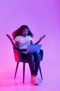 Genç siyah bir kadın, dizüstü bilgisayarında çalışırken şok olmuş bir şekilde koltukta oturuyor. Neon ışıkları, iş projesindeki bir hatadan dolayı şaşkınlığını ifade ederken canlı bir atmosfer yaratıyor..