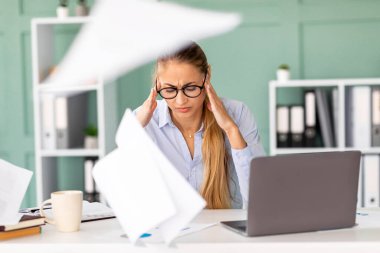 Yoğun bir ofiste masasında oturan bir kadın şakaklarına dokunuyor ve stresli görünüyor. Gazeteler havada uçuşuyor. Yorgun ve iş yoğunluğundan bunalmış görünüyor. Rutin işlerle boğuşuyor..