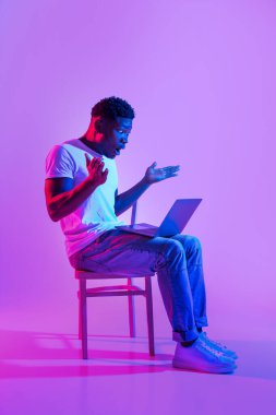 Genç siyahi bir adam bir sandalyede oturuyor, dizüstü bilgisayar kullanırken şaşkın ve sıkıntılı görünüyor. Çevrimiçi bir iş projesinin son teslim tarihinin stresiyle yüzleşirken, neon ışığı dramatik bir atmosfer yaratıyor..