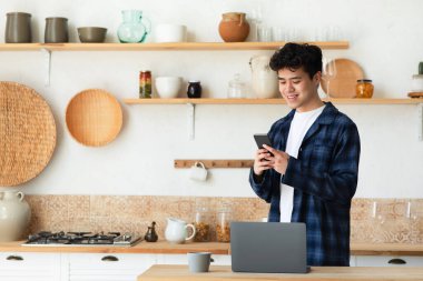 COVID-19 salgını ve sosyal uzaklık sırasında evde çalışma ve eğitim için modern cihazlar. Gülümseyen milenyum Asyalı erkek modern mutfak mutfağında dizüstü bilgisayar ve bardakla telefonda yazıyor.