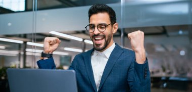 Genç bir adam dizüstü bilgisayarın önünde otururken heyecan ve mutluluk gösteriyor. Bir amaca ulaştığını ya da bir görevi tamamladığını öne sürerek kutlama için kollarını kaldırır..
