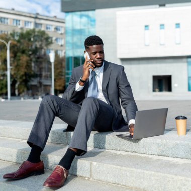 Kendine güvenen Afro-Amerikan iş adamı müşterileriyle cep telefonuyla konuşuyor ve dışarıda dizüstü bilgisayar kullanıyor, ofis binasına karşı oturuyor, ürün topluyor. Ofis dışında çalış.