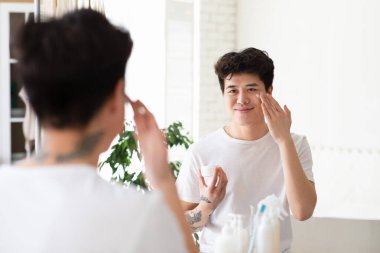 Nemlendirici cilt, güzellik ve günlük işlemleri evde kozmetik ürünlerle yapmak. Mutlu Asyalı genç adam yüzüne krem sürüyor ve banyo içindeki ayna yansımasına bakıyor, boşluğu kopyalıyor.
