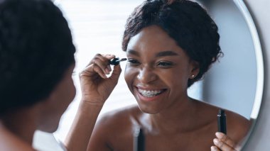 Bir kadın, aynanın önünde kirpiklerine rimel sürerken gülümsüyor. Oda aydınlık ve makyajını tazelerken odaklandığını gösteriyor. Bu an günlük rutini yakalar..