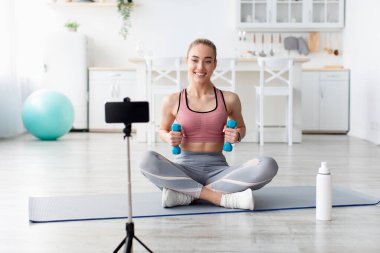 Neşeli Avrupalı bin yıllık sarışın fitness blogcusu webcam 'e bakar ve evde sosyal ağlar için halterli antreman videosu çeker. Modern blog, spor ve aygıt, yeni normal ve güvenli