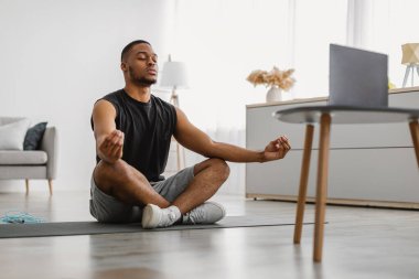 Çevrimiçi yoga. Huzurlu Afrikalı Amerikalı Adam Lotus pozisyonunda oturmuş evde meditasyon videosu izleyerek meditasyon yapıyor. Meditasyon Rahatlatıcı Uygulama ve Egzersiz