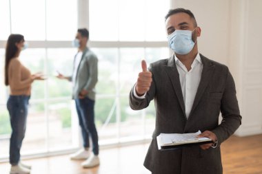 Gayrimenkul, Satış ve İnsanlar Konsepti. Yüz maskesi takmış mutlu, gülümseyen profesyonel satıcının portresi. Elinde not panosu var. Baş parmak işareti, seçici odaklanma. Müşteriler satın almayı tartışıyor