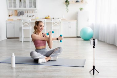 Yeni normal, evde fitness dersleri, online antrenman ve uzaktan antrenörlük. Gülümseyen sarışın bayan paspasın üzerine oturmuş dambıllarla egzersiz yapıyor oturma odasındaki internet kameralarına şişeyle bakıyor.