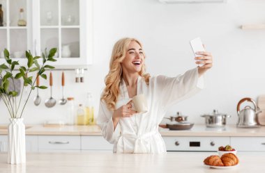 Cüppeli, güzel, Avrupalı, genç, sarışın bir kadın kahve içiyor, kahvaltıda, İskandinav mutfağının iç kısımlarında akıllı telefondan selfie çekiyor, fotokopi çekiyor. Modern aygıt, uygulama, blog ve sosyal ağlar
