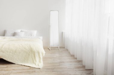 Yeni tasarım yenileme veya yeniden yerleştirme, emlak reklamı ve blog. Yastıklı yatak, gün ışığında ahşap zeminde halı perdeli, aynalı, yatak odasının arka planında beyaz bir duvar var.