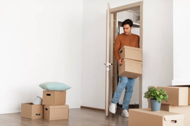 Genç bir milenyum adamı kapı aralığından geçiyor, karton kutuları dengeliyor. Yeni dairesine taşınırken odaklanmış ve meşgul görünüyor. Yer boş ve ışık dolu..