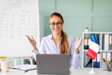 Genç bir bayan öğretmen, dizüstü bilgisayarına bakarken kuralları açıklayan çevrimiçi Fransızca dersi veriyor. Öğretim web konferansı sırasında öğrencilerle etkileşim içinde, öğretmeye hevesli..